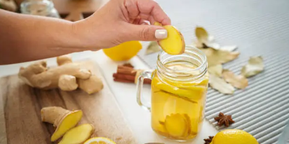 Main plaçant une tranche de gingembre dans un verre de jus de citron au gingembre, entouré de morceaux de gingembre frais et de citrons sur une table, idéal pour une boisson détoxifiante et énergisante.